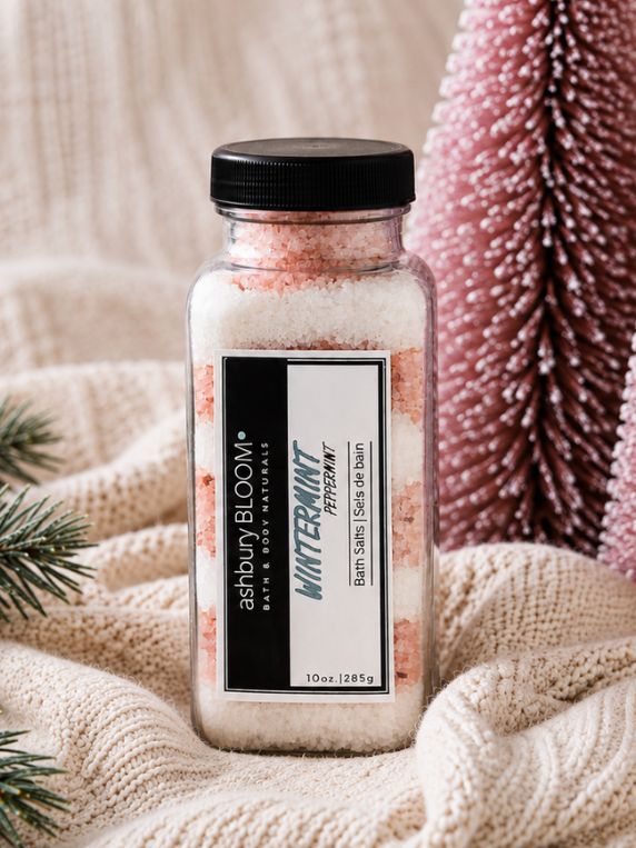 Ashbury Bloom Wintermint Peppermint Bath Salts in a clear jar with pink and white bath salts, displayed on a cream knit blanket with evergreen branches and pink Christmas trees.