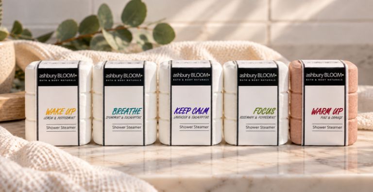 Five ashbury BLOOM• shower steamer varieties—Wake Up, Breathe, Keep Calm, Focus, and Warm Up—displayed on a marble surface with a cream towel and eucalyptus.