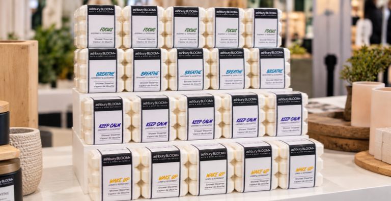 Ashbury BLOOM• shower steamers arranged in rows on a tiered white display stand at a wholesale trade show, showcasing the packaged collection.